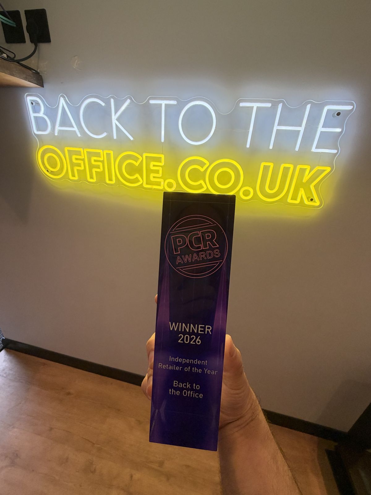 The PCR Awards 2026 trophy for Independent Retailer of the Year, held up in front of the illuminated Back to the Office sign